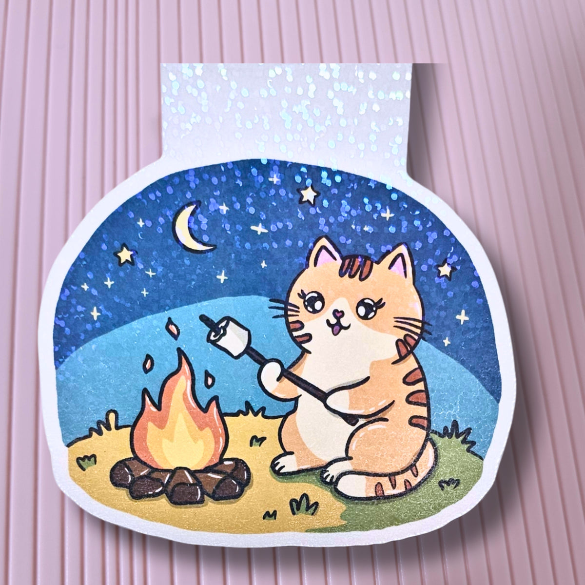 Lesezeichen mit dem Motiv von Mieze Theo, der sitzt am Lagerfeuer und brät sich Marshmallows. Über ihm Sternenhimmel und Mond.